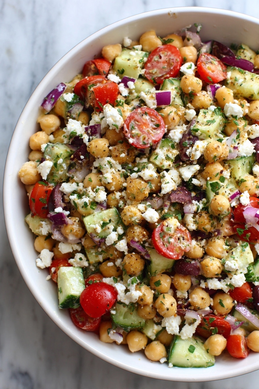 The image shows a close-up of a Greek-style chickpea salad in a white bowl. The salad layers include light tan chickpeas spread out evenly, mixed with bright red cherry tomato halves, green celery pieces, chopped cucumber, small dark purple olive pieces, and small bits of red onion. White crumbled feta cheese is sprinkled generously on top, giving a textured, crumbly look. Tiny green herbs are scattered throughout the salad for extra color. Everything is mixed together with visible creamy dressing that lightly coats the ingredients. The background is a white marbled surface. Photo taken with an iphone --ar 2:3 --v 7 - Healthy Chickpea Tuna Salad, healthy tuna salad, protein-packed lunch, easy seafood salad, nutritious chickpea salad