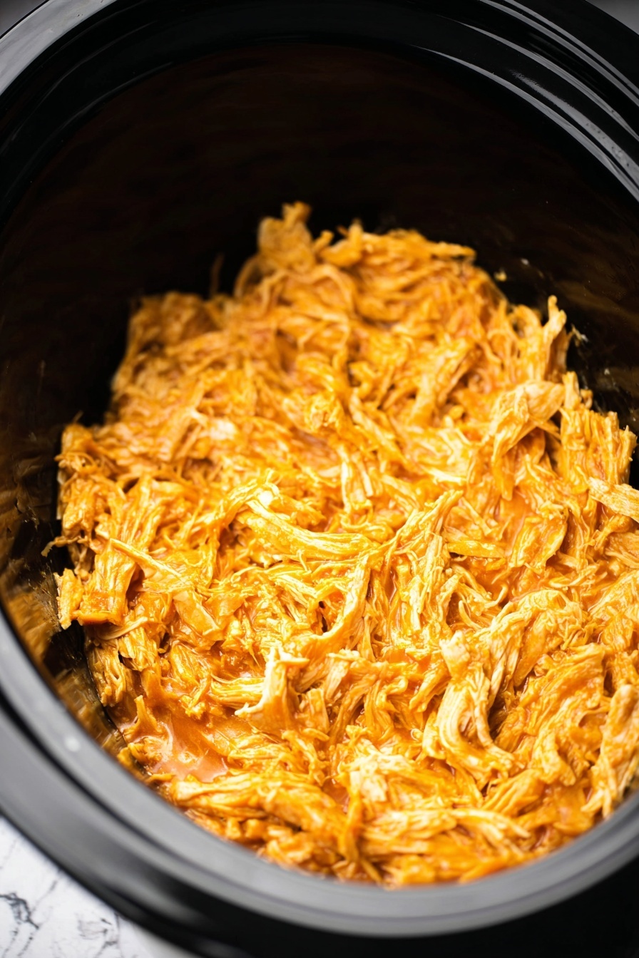 A close-up view of shredded orange chicken inside a black slow cooker. The chicken is finely shredded with moist, tender strands, coated evenly in a bright orange sauce that gives a slightly glossy texture. The black slow cooker bowl has a smooth, shiny surface, and the shredded chicken fills the bottom half of the cooker, showing a mix of smaller and larger pieces. The background is a white marbled texture photo taken with an iphone --ar 2:3 --v 7 - Slow Cooker Buffalo Chicken, Buffalo Chicken Recipes, Easy Slow Cooker Chicken, Spicy Chicken Dinner, Make-Ahead Chicken Recipes