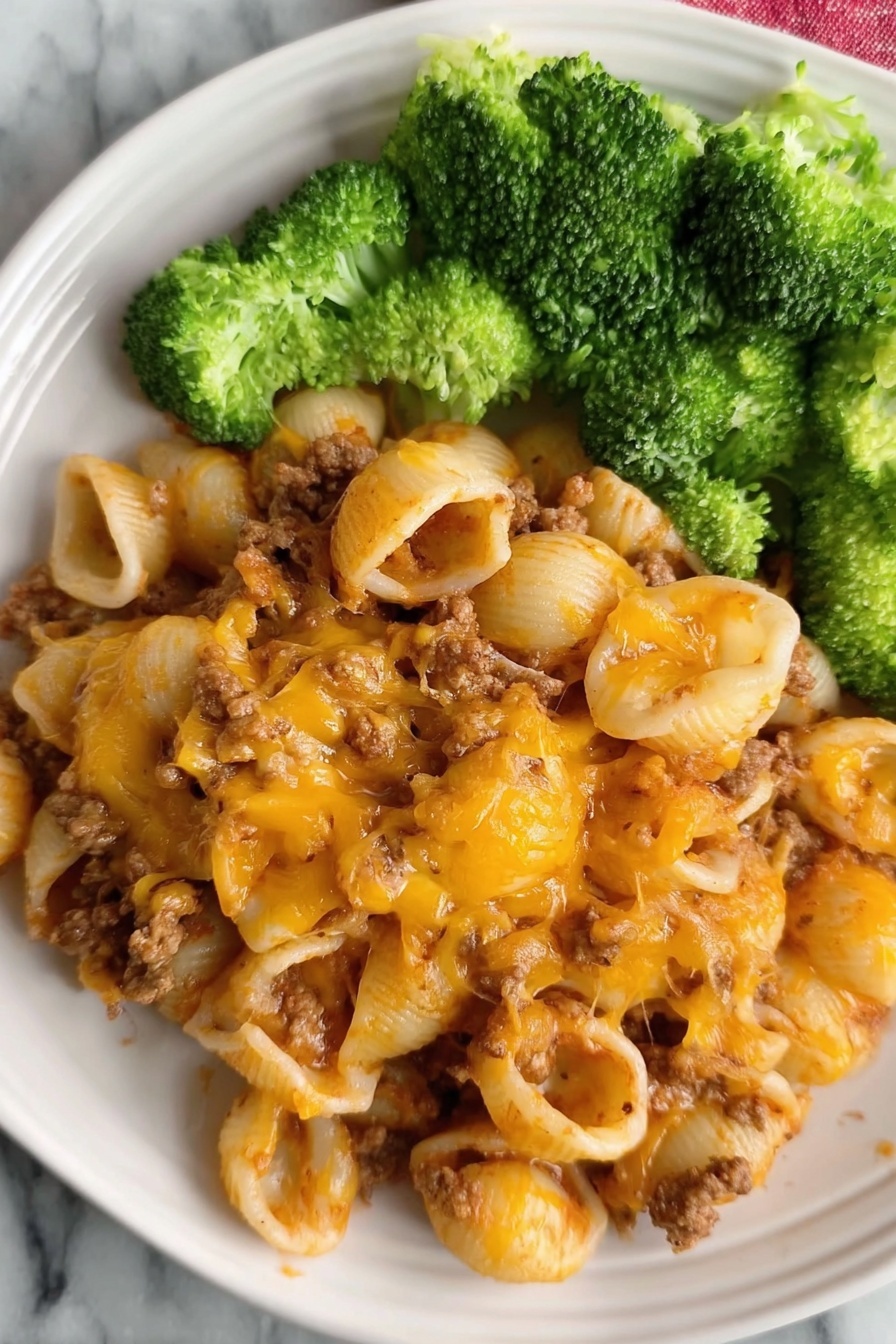 The dish shows a plate filled with three layers: the bottom layer is small pasta shells mixed with cooked ground meat that is soft brown in color; the middle layer is melted cheddar cheese with an orange and golden-brown texture spread evenly over the pasta, giving it a slightly crispy look in places; the top layer is bright green broccoli florets placed on one side of the plate. The plate is white, and the surface beneath is white marble. Photo taken with an iphone --ar 2:3 --v 7 - Easy Hamburger Pasta Casserole, cheeseburger pasta bake, quick pasta casserole, ground beef pasta recipe, family-friendly dinner ideas