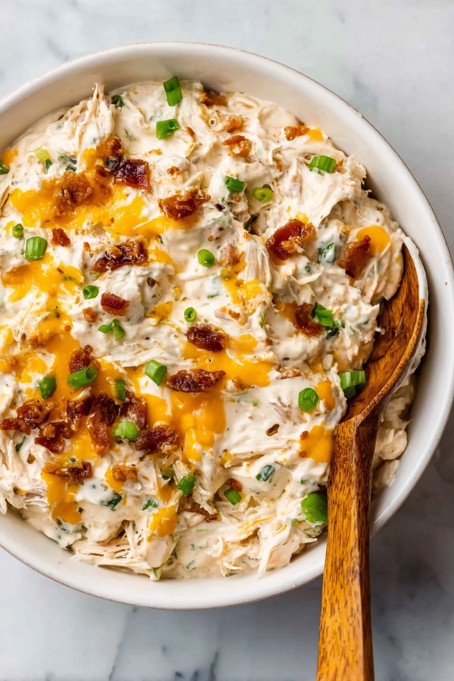 The image shows a white bowl filled with a creamy dip that has a thick, chunky texture. The dip contains visible strands of shredded white meat mixed with creamy white sauce. There are small bright orange melted cheese drops spread over the top and small pieces of green onion scattered throughout. Crispy brown bits, likely bacon or a similar garnish, are also mixed in, adding texture. A wooden spoon with a smooth surface rests inside the bowl on the right side. The bowl is placed on a white marbled surface. photo taken with an iphone --ar 2:3 --v 7 - Crock Pot Crack Chicken, creamy slow cooker chicken, shredded chicken recipes, easy comfort food, bacon chicken dinner