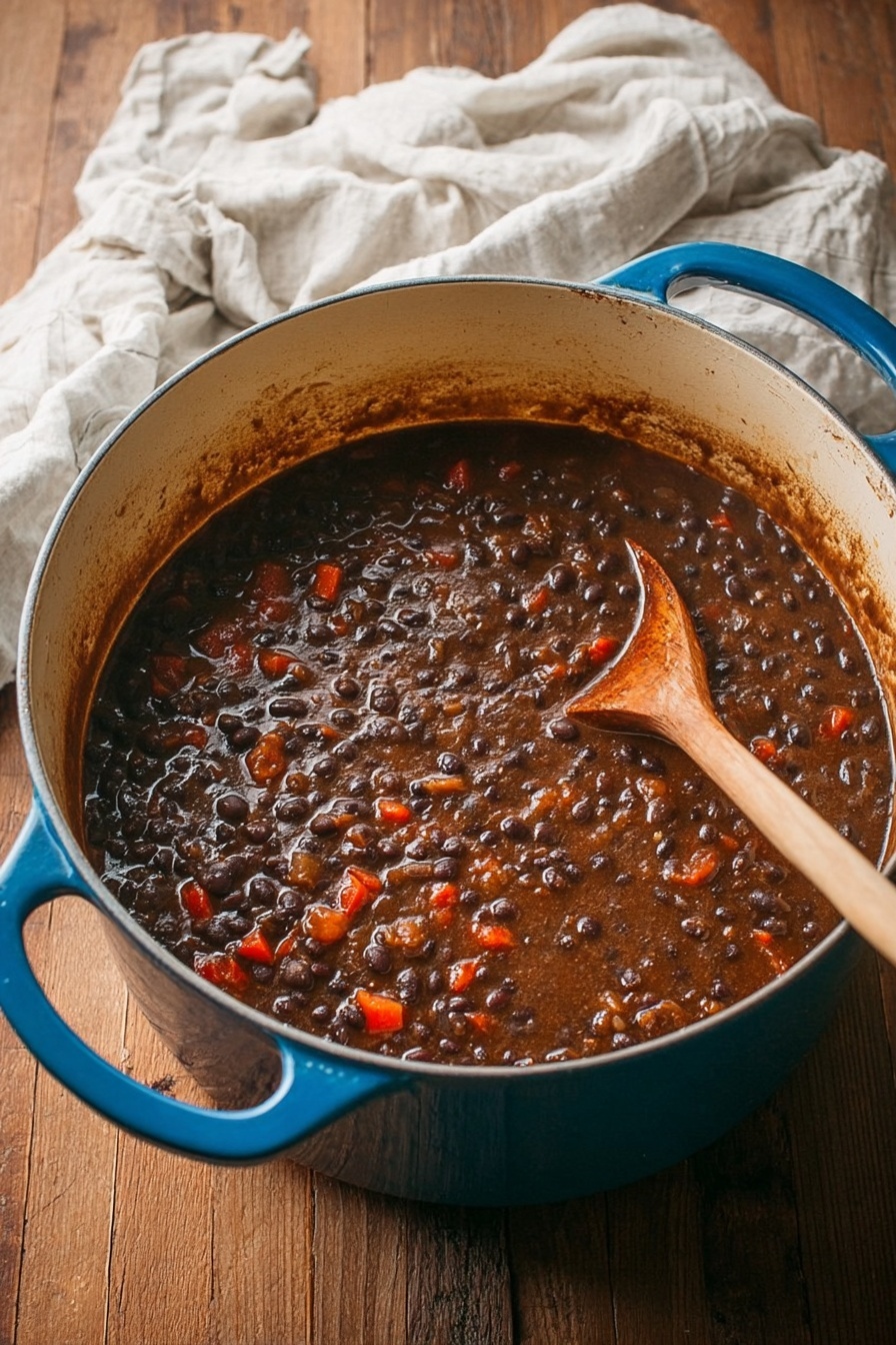 Spicy Black Bean Soup Recipe 6 A large blue pot filled with thick dark brown stew, full of black beans and small diced orange-red pieces that look like bell peppers or carrots, the stew has a rich, slightly chunky texture. Inside the pot, there is a wooden spoon resting on the right side, partially covered by the stew. The pot sits on a light wooden table with a crumpled off-white cloth near the top edge. The photo taken with an iphone --ar 2:3 --v 7 - Spicy Black Bean Soup, black bean soup recipes, easy spicy soup, hearty vegetarian soup, smoky bean soup