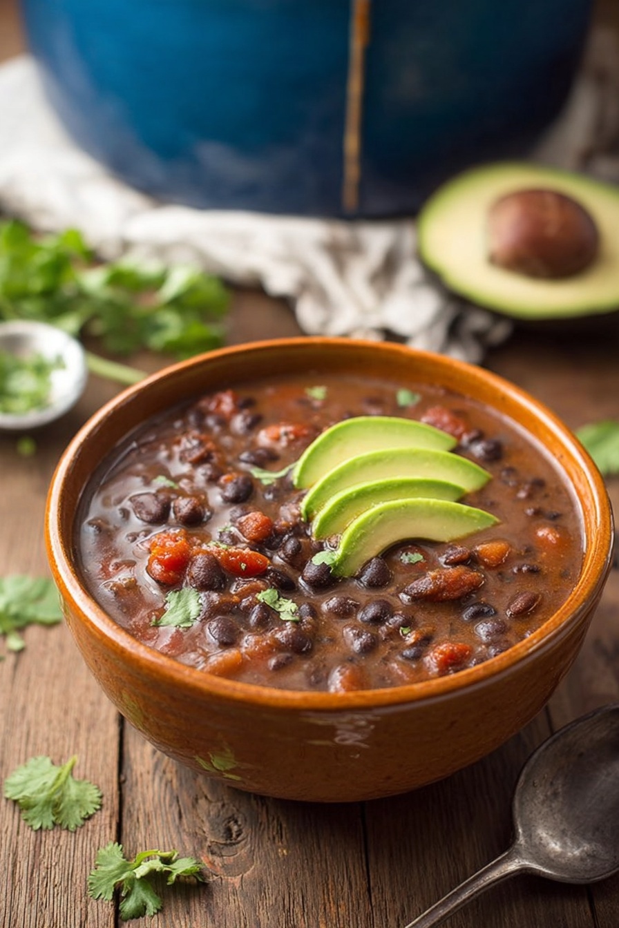 Spicy Black Bean Soup Recipe 8 A brown bowl filled with thick black bean soup shows dark black beans and small pieces of reddish tomatoes mixed in a rich brown broth. On top, there are three thin slices of green avocado stacked on the right side and some bright green cilantro leaves on the left side. The bowl sits on a wooden surface with loose cilantro leaves scattered nearby. Behind the bowl, a half avocado with a brown seed rests on a white cloth. A large blue pot of the same soup is blurred in the background, with a drip down its side. A metal spoon lies to the right of the bowl. Photo taken with an iphone --ar 2:3 --v 7 - Spicy Black Bean Soup, black bean soup recipes, easy spicy soup, hearty vegetarian soup, smoky bean soup