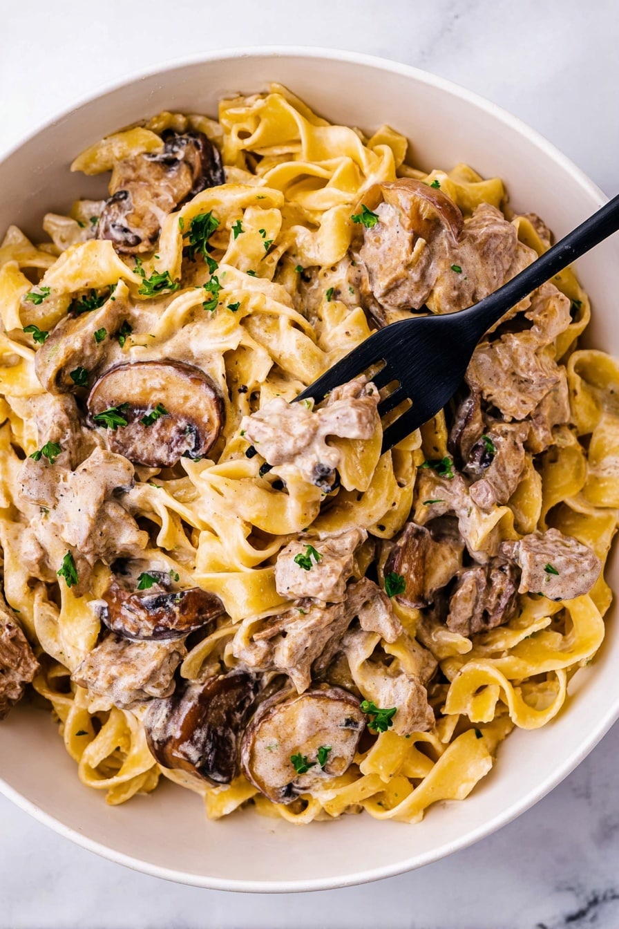 A white bowl filled with a creamy pasta dish made of multiple layers: the base layer is yellow egg noodles with a smooth texture, mixed evenly throughout are light brown strips of cooked meat, and slices of dark brown mushrooms coated in a creamy light beige sauce. Small green parsley pieces are scattered on top, adding color contrast. A black fork holds a piece of meat and a mushroom slice in the center of the bowl. The bowl is placed on a white marbled surface photo taken with an iphone --ar 2:3 --v 7 - Easy Beef Stroganoff, beef stroganoff recipe, quick beef dinner, creamy beef skillet, Russian-inspired beef dish