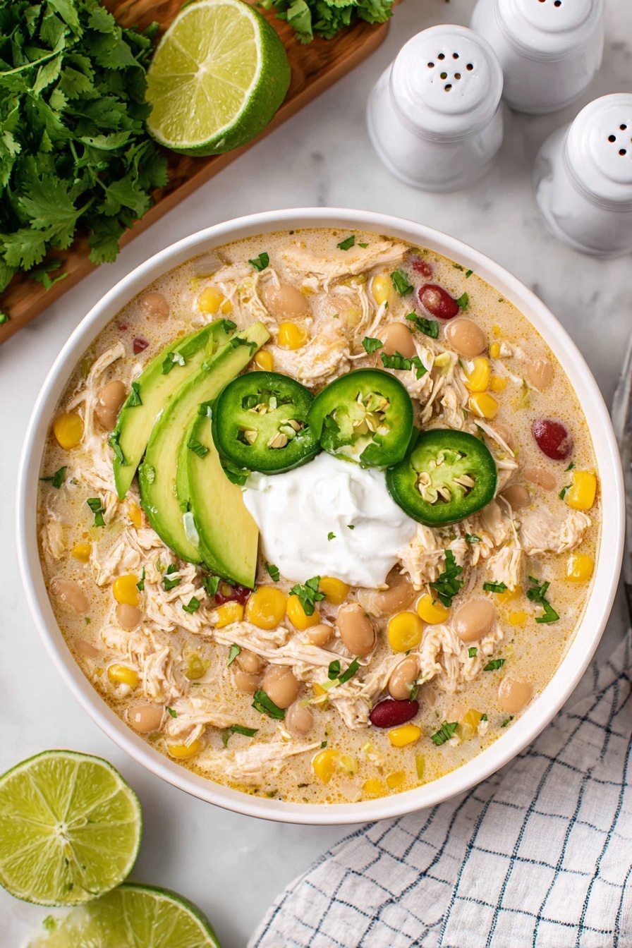 Creamy Crockpot White Chicken Chili Recipe 8 A white bowl filled with creamy chicken soup with visible shredded chicken, white and red beans, and yellow corn kernels spread throughout. There are three slices of green jalapeño placed on top, alongside three smooth green avocado slices arranged on one edge. In the center sits a dollop of white sour cream. Two thin lime wedges rest on the side of the bowl. The bowl is on a white marbled surface with green cilantro and lime on a wooden board nearby, and salt and pepper shakers in the background. A white and blue checkered cloth is placed beside the bowl. photo taken with an iphone --ar 2:3 --v 7 - Creamy Crockpot White Chicken Chili, White Chicken Chili Crockpot, Easy White Chicken Chili, Crockpot Chicken Chili, White Chicken Chili Recipe