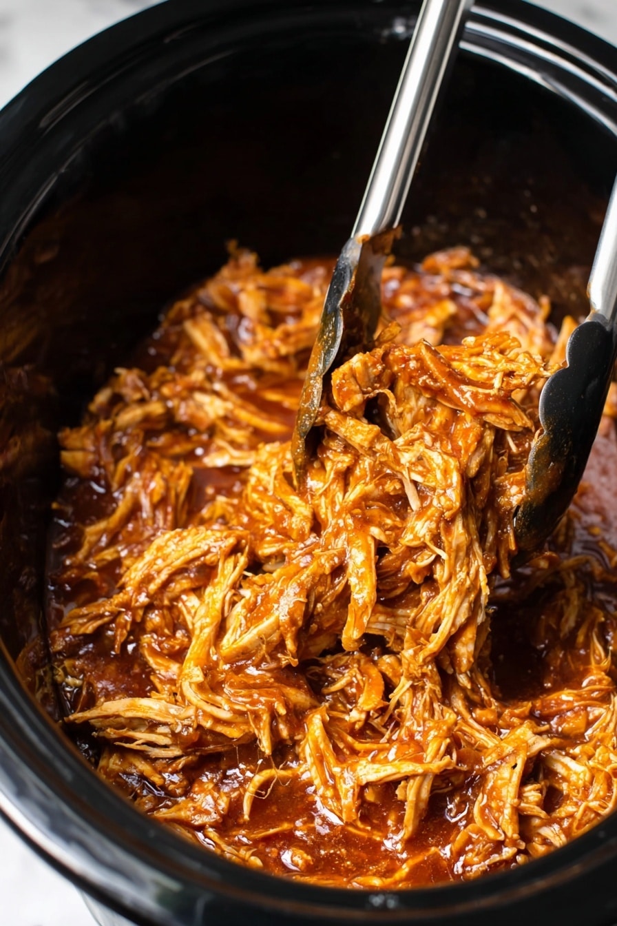 The image shows a close-up of shredded chicken in a slow cooker, mixed well with a rich, glossy, reddish-brown sauce. The chicken pieces have a tender texture and are coated evenly by the thick sauce. A pair of silver and black tongs lifts some of the sauced chicken from the slow cooker, showing the mix of small shredded parts and slightly bigger chunks. The background and edges of the slow cooker are black, contrasting with the warm colors of the chicken dish. The photo is set on a white marbled surface. photo taken with an iphone --ar 2:3 --v 7 - Crockpot BBQ Chicken, easy slow cooker chicken, flavorful crockpot chicken recipes, tender BBQ chicken, simple dinner ideas