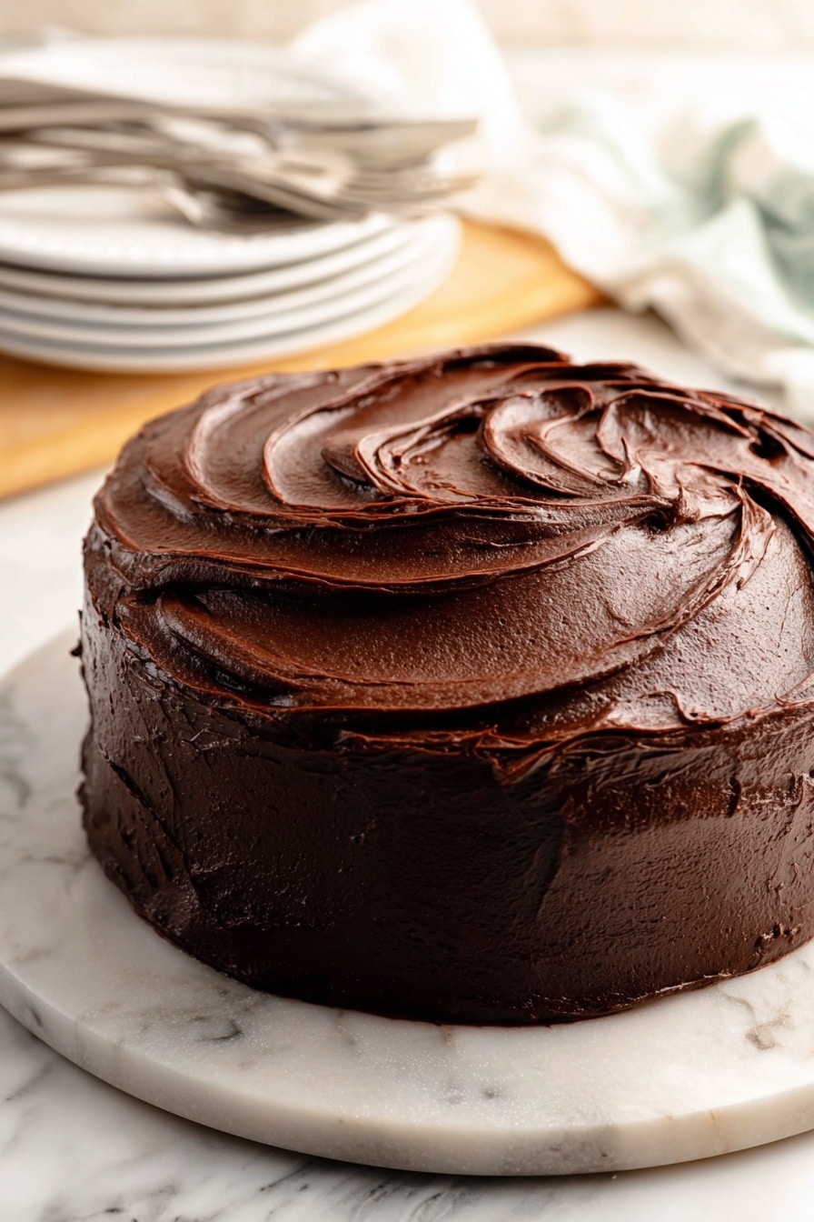 Decadent Chocolate Cake with Fudge Frosting Recipe 8 A round chocolate cake with a thick, smooth, and glossy dark brown frosting covering the whole cake. The frosting is spread in large, curved swirls and waves, giving it a rich and creamy texture. The cake sits on a white marbled round board. In the background, there are stacked white plates with ridged edges and silver forks on a white marbled surface. The photo taken with an iphone --ar 2:3 --v 7 - Decadent Chocolate Cake with Fudge Frosting, rich chocolate cake recipe, moist chocolate cake with fudge frosting, easy chocolate cake dessert, chocolate cake with luscious frosting