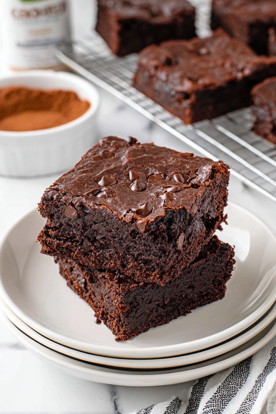Cottage Cheese Brownies with Chocolate Chips Recipe 6 Two thick, dark brown chocolate brownies are stacked on a set of white plates, showing a rich, dense texture with visible chocolate chips embedded throughout the top layer. The brownies have a slightly crumbly edge with a moist interior. In the background, more brownies rest on a cooling rack, and a container of cocoa powder is partially visible. The setting includes a white marbled surface and a striped cloth nearby. Photo taken with an iphone --ar 2:3 --v 7 - Cottage Cheese Brownies with Chocolate Chips, healthy chocolate chip brownies, moist fudge brownies, creamy cottage cheese dessert, easy brownie recipes