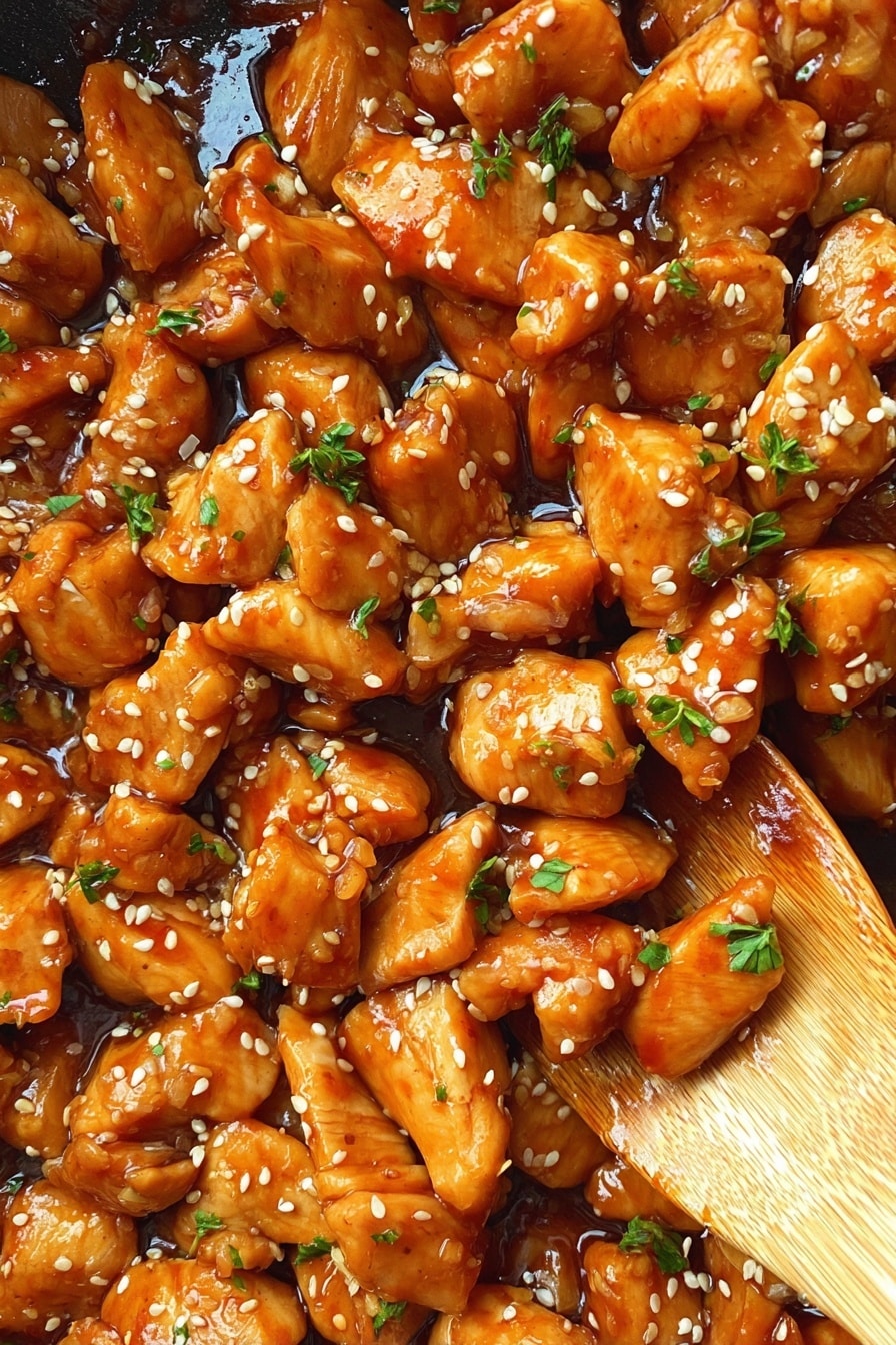 Easy Teriyaki Chicken Bowl Recipe 8 A close-up of a dish with many pieces of cooked chicken, all coated in a shiny, thick orange-brown sauce. The pieces are cut into small, irregular shapes with a smooth, moist texture. White sesame seeds are sprinkled evenly across the chicken, along with small bits of bright green herbs for contrast. A wooden spoon is partially visible in the bottom right part of the image, resting lightly on some of the chicken. The background shows a glossy surface of the sauce underneath the chicken. photo taken with an iphone --ar 2:3 --v 7 - Easy Teriyaki Chicken Bowl, easy chicken bowl, homemade teriyaki sauce, quick dinner recipe, healthy rice bowl