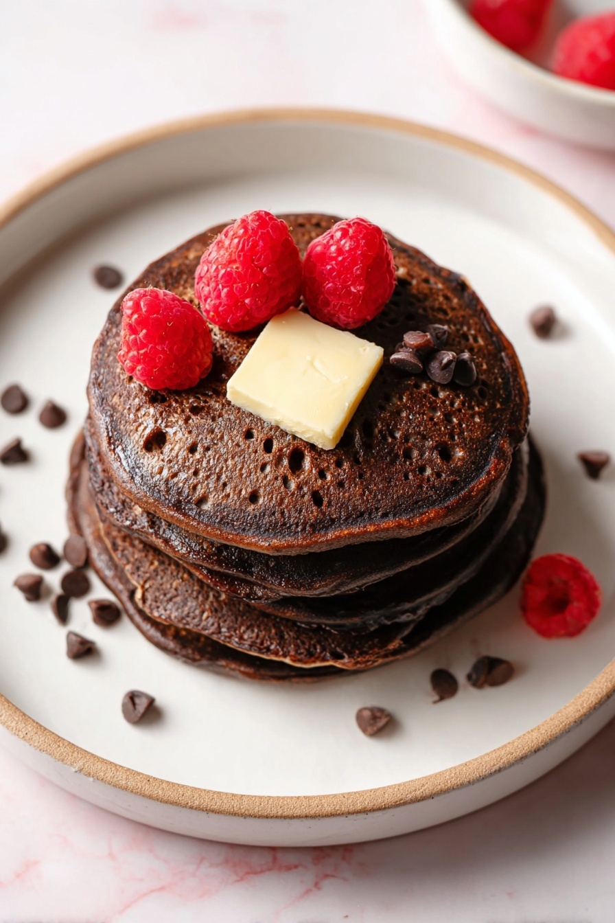 Chocolate Pancakes with Melting Chocolate Chips Recipe 8 A stack of four dark brown chocolate pancakes is placed on a white plate with a thin beige rim, set on a white marbled surface. On the top pancake, there is a square piece of light yellow butter in the center. Three bright red raspberries are arranged around the butter, and small dark brown chocolate chips are scattered on top of the butter and on the plate near the stack. The pancakes have a slightly porous texture with small holes visible. Photo taken with an iphone --ar 2:3 --v 7 - Chocolate Pancakes with Melting Chocolate Chips, decadent chocolate pancakes, fluffy chocolate breakfast, easy chocolate pancake recipe, breakfast with chocolate chips
