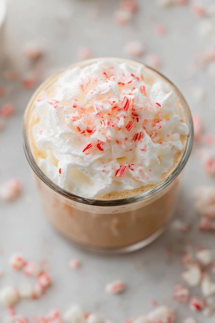 Peppermint White Mocha Recipe 8 A clear glass filled with a drink that has a light brown layer at the bottom, topped with a thick, fluffy white whipped cream layer. On top of the whipped cream, there are small red and white crushed candy pieces spread evenly. The glass sits on a white marbled surface with blurry white and red candy pieces scattered around. The photo is taken from above, showing mostly the whipped cream and candy topping. photo taken with an iphone --ar 2:3 --v 7 - Peppermint White Mocha, holiday peppermint mocha, festive coffee drinks, peppermint hot chocolate, white chocolate peppermint beverage
