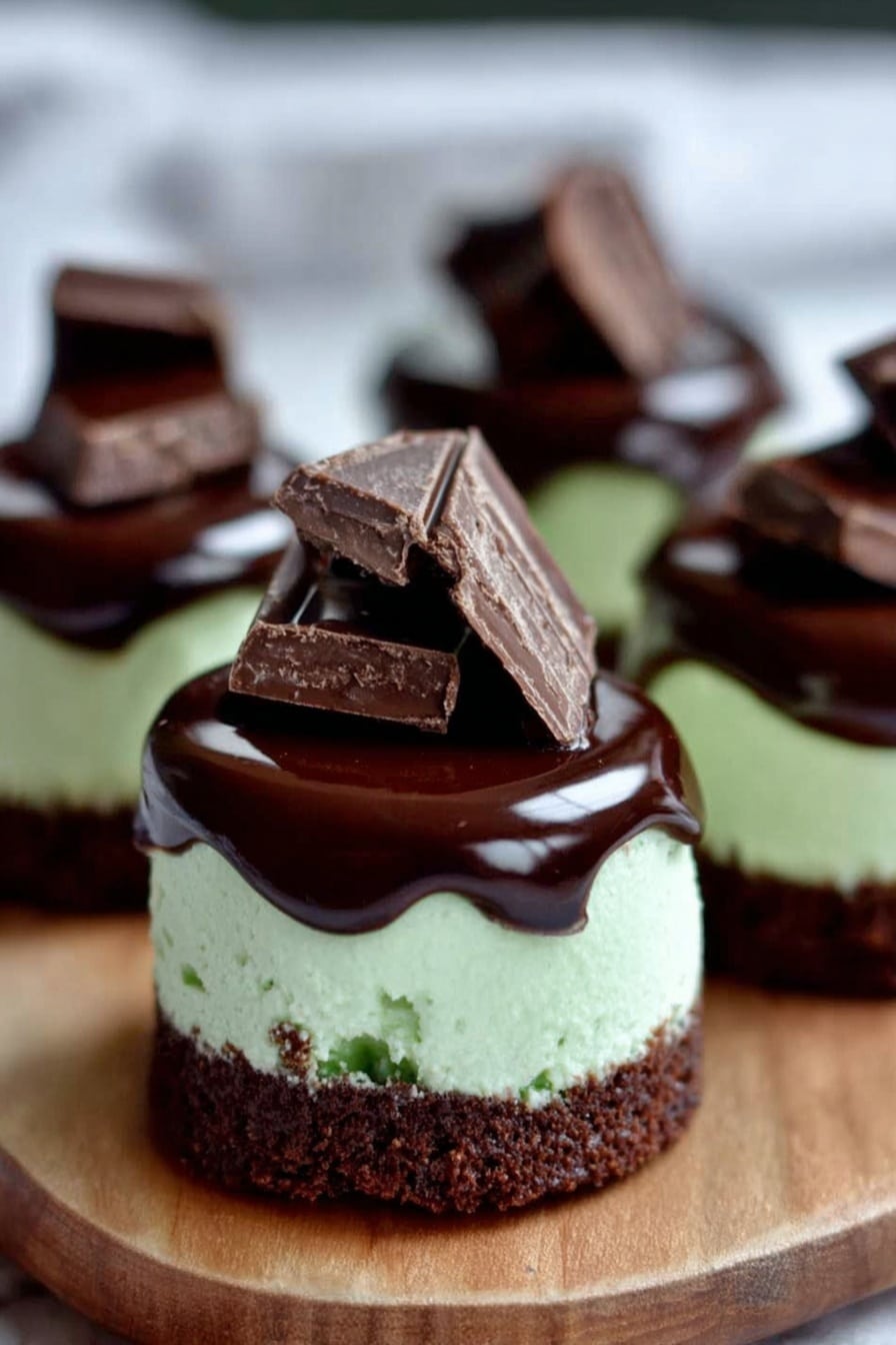 Andes Mint Mini Cheesecakes Recipe 6 The image shows mini cakes with three layers. The bottom layer is dark brown and looks like a crumbly chocolate base. The middle layer is a light green, smooth, and creamy mint filling. The top layer is a shiny dark chocolate glaze that spreads a little down the sides. On top of each cake, there are two small pieces of dark chocolate stacked unevenly. The cakes are placed on a wooden board with a white marbled texture in the background. Photo taken with an iphone --ar 2:3 --v 7 - Andes Mint Mini Cheesecakes, mint chocolate cheesecake bites, mini cheesecakes with Andes mints, no-bake mint cheesecakes, holiday cheesecake desserts
