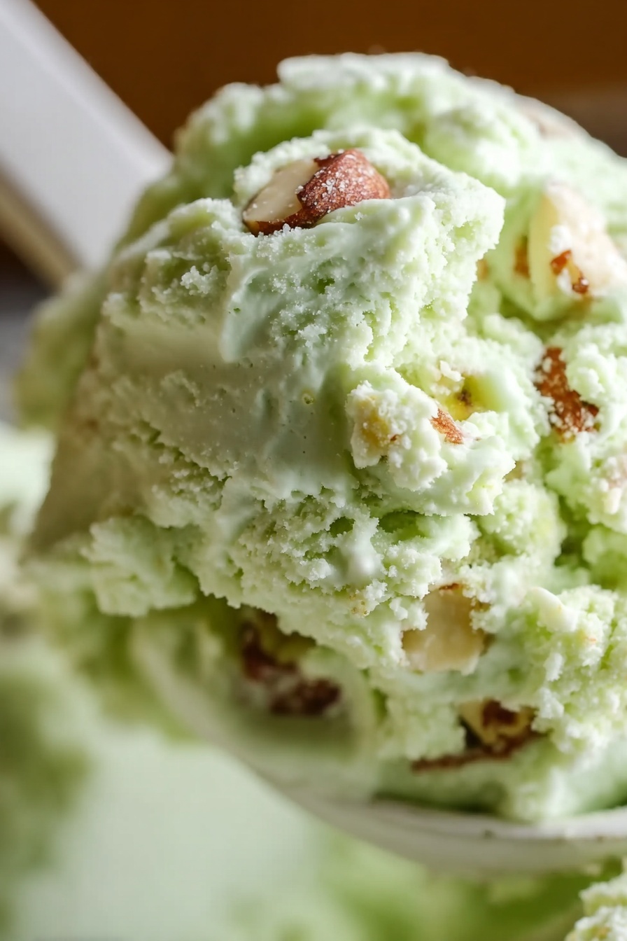 Pistachio Pudding Cookies Recipe 8 This close-up image shows a scoop of light green ice cream with a creamy texture. Mixed throughout the ice cream are small white chunks and pieces of nuts, which add a slight crunchy texture. The ice cream is scooped on a white spoon, with the background softly blurred and featuring a warm brown tone. The focus is on the mix of smooth ice cream and the chunks within, making it look rich and inviting. photo taken with an iphone --ar 2:3 --v 7 - Pistachio Pudding Cookies, pistachio cookies with pudding mix, chewy pistachio cookies, white chocolate pistachio cookies, easy pistachio cookie recipe