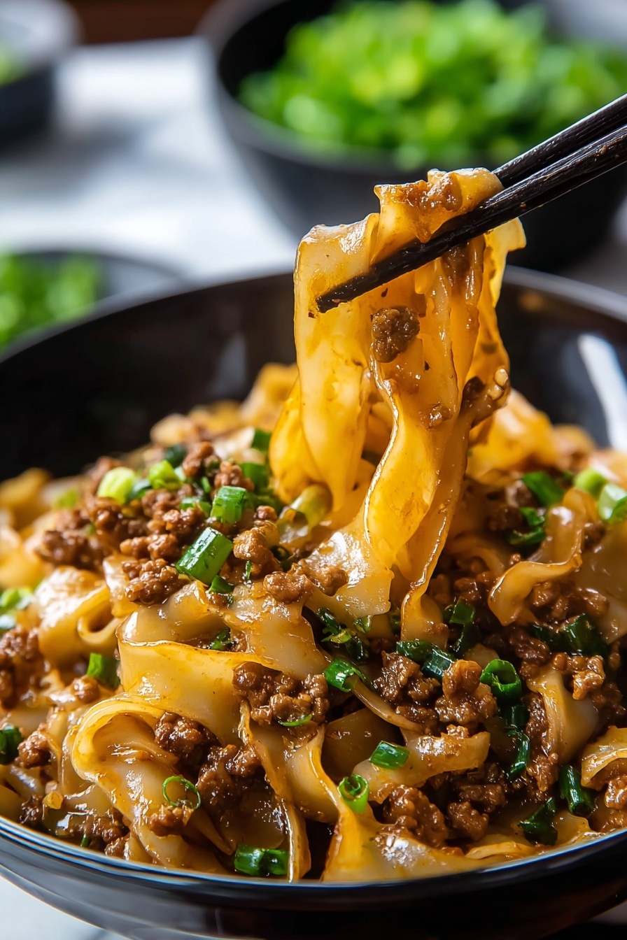Potsticker Noodle Bowl with Pork and Greens Recipe 6 A close-up view of wide flat noodles mixed with small brown pieces of ground meat and green chopped chives, all covered in a shiny brown sauce. The noodles sit in a black bowl on a white marbled surface, with a pair of black chopsticks lifting a clump of noodles and meat from the bowl. In the blurred background, there is a round black bowl filled with more green chopped chives. The scene looks warm and savory, showing texture in the noodles and meat with a glossy finish. Photo taken with an iphone --ar 2:3 --v 7 - Potsticker Noodle Bowl with Pork and Greens, Asian noodle bowl recipes, quick weeknight dinners, savory pork noodle dishes, healthy Asian dinner ideas