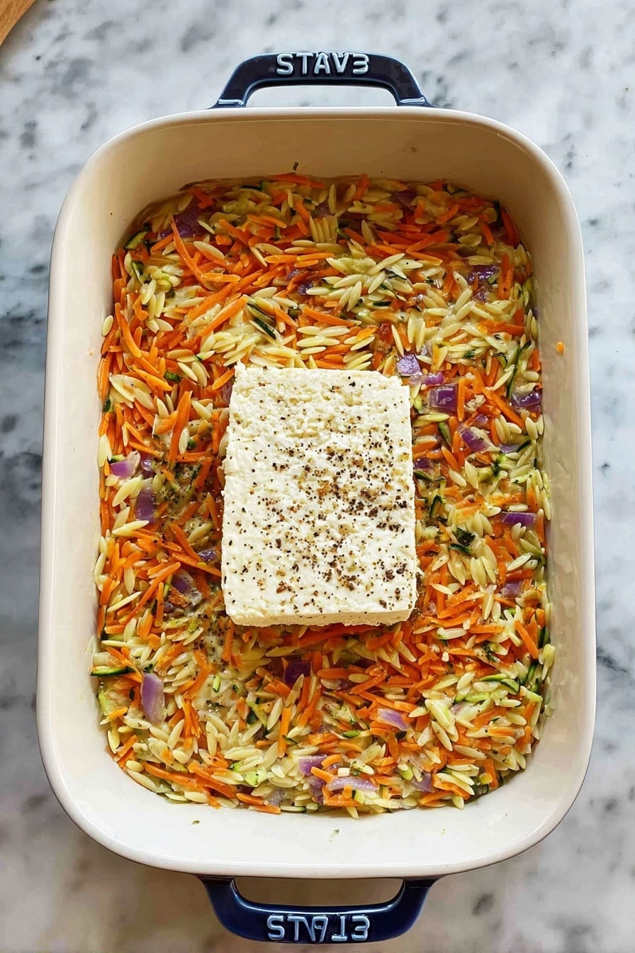 A white rectangular baking dish filled with a layered mixture of shredded orange carrots, small orange orzo pasta, grated green zucchini, and small purple onions, all mixed in a creamy base. In the center of the dish, there is a single thick square of white cheese topped with black pepper. The baking dish has a blue handle with the brand name