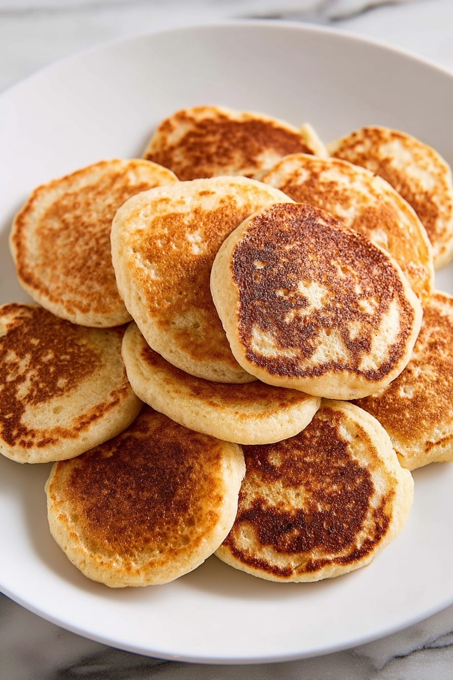 A white plate holds a stack of eight small, round pancakes arranged in a loose pile. Each pancake has a light brown and golden color with darker, slightly crisp spots on the surface, showing a mix of soft and cooked textures. The edges are smooth and slightly raised, and the pancakes are fluffy with small air holes visible. The plate sits on a white marbled surface with natural light highlighting the warm tones of the pancakes. photo taken with an iphone --ar 2:3 --v 7 - Banana Oatmeal Pancakes, healthy breakfast recipes, quick pancake ideas, wholesome banana pancakes, easy oat pancake recipe