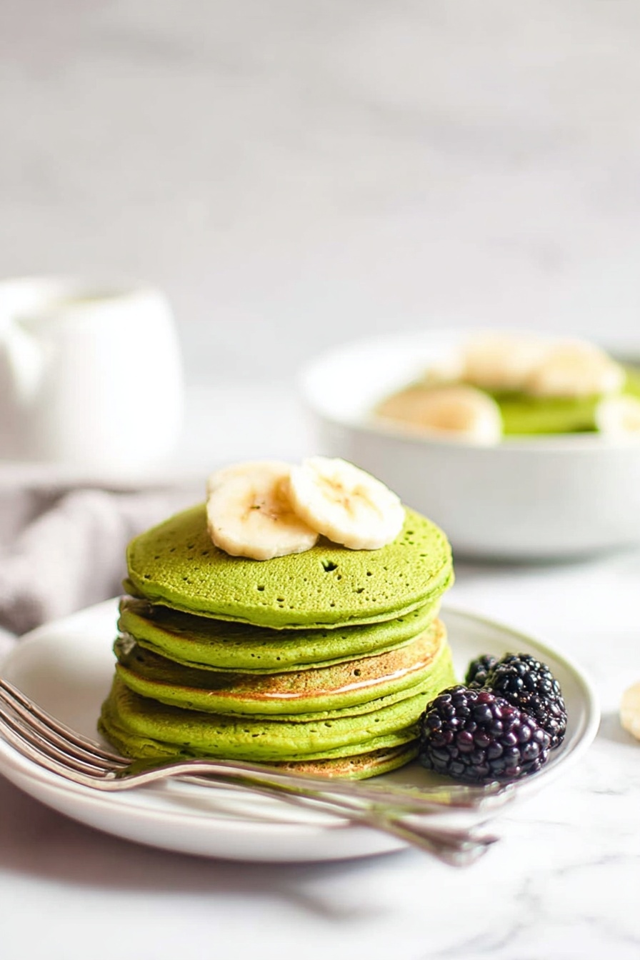 Banana Spinach Blender Pancakes Recipe 8 A stack of five green pancakes is placed in the center of a white plate on a white marbled surface. The pancakes have a smooth texture with small holes visible on the sides. On top of the stack, there are three slices of banana. Two blackberries rest next to the pancakes on the plate. To the right side, a silver fork and knife are positioned beside the stack. In the background, a white bowl with more green pancakes and banana slices is slightly blurred. The overall scene is bright with soft lighting. photo taken with an iphone --ar 2:3 --v 7 - Banana Spinach Blender Pancakes, healthy green pancake recipe, quick nutritious breakfast, easy spinach banana pancakes, fluffy green pancake recipe