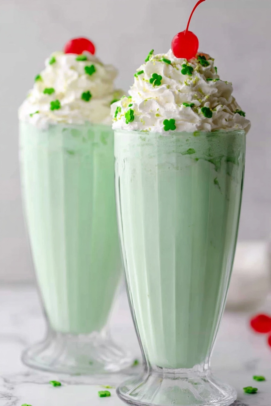 Mint Shamrock Shake Recipe 6 Two tall, clear glasses filled with light green milkshake that looks smooth and creamy stand side by side on a white marbled surface. Each glass has a generous swirl of white whipped cream on top, decorated with small green clover-shaped sprinkles scattered around and a bright red cherry placed at the peak. The light green milkshake is thick and fills most of the glass, which has a ridged pattern that adds texture. The background is simple and softly blurred, keeping the focus on the milkshake glasses. Photo taken with an iphone --ar 2:3 --v 7 - Mint Shamrock Shake Mint Shamrock Shake Recipe St. Patrick's Day Mint Milkshake