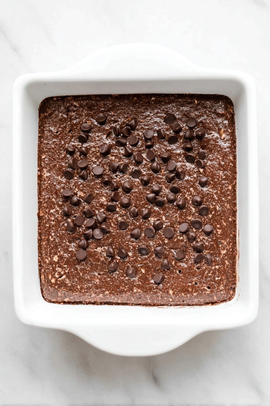 Cottage Cheese Brownies with Chocolate Chips Recipe 8 A square white baking dish filled with a thick, chocolatey mixture that is dark brown in color with small oat-like texture spots throughout. The top is covered with scattered dark chocolate chips that are slightly shiny and sit unevenly on the surface. The surface looks dense and slightly rough, with some small cracks visible near the edges. The dish is placed on a white marbled texture background. photo taken with an iphone --ar 2:3 --v 7 - Cottage Cheese Brownies with Chocolate Chips, healthy chocolate chip brownies, moist fudge brownies, creamy cottage cheese dessert, easy brownie recipes