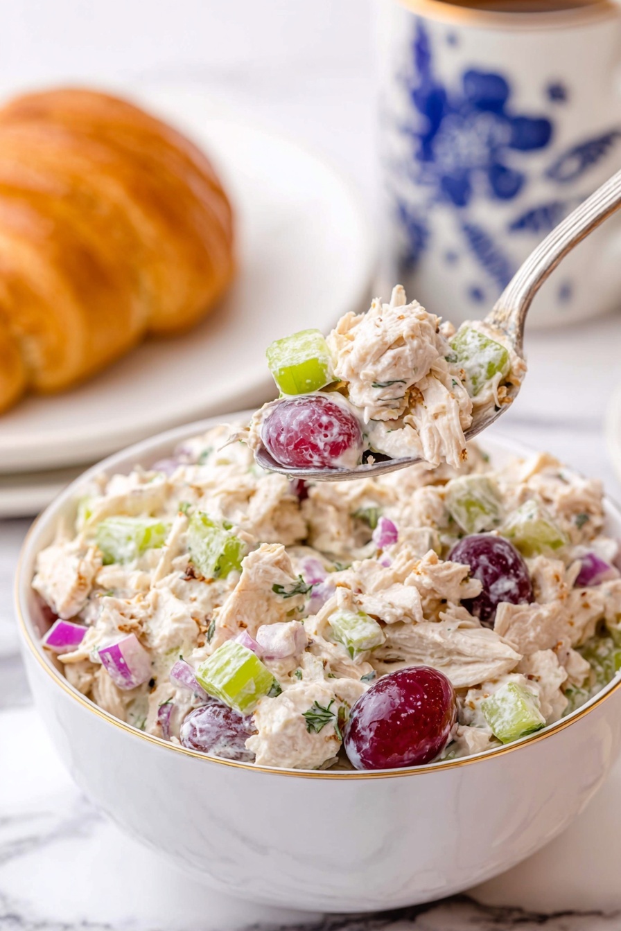 A white bowl filled with a creamy chicken salad showing shredded light-colored chicken mixed with light green celery pieces, small purple-red grape halves, small bright purple onion chunks, and some specks of herbs. A silver spoon is lifting a spoonful of the chicken salad above the bowl, highlighting the colorful mix. In the background, there is a white cup with blue floral designs and a white plate holding a golden croissant, all placed on a white marbled surface. photo taken with an iphone --ar 2:3 --v 7 - Grapes Chicken Salad, easy chicken salad, fruit chicken salad, healthy lunch recipes, quick salad ideas