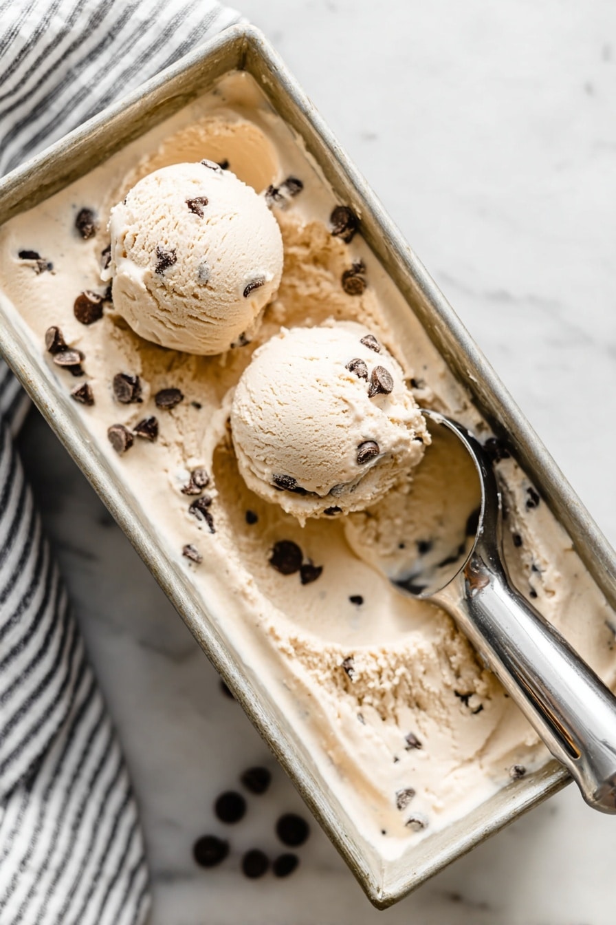 Creamy Cottage Cheese Ice Cream with Chocolate Recipe 6 The image shows a rectangular metal loaf pan filled with light tan ice cream mixed with dark chocolate chips. On top of the ice cream, there are two perfectly round scoops, one closer to the front left and the other near the middle right, both containing visible chocolate chips. A metallic ice cream scooper holding some ice cream rests on the right side of the pan. The pan is placed on a white marbled surface, and a striped cloth is faintly visible in the top left corner. The overall scene looks soft and creamy with a cool, fresh feeling. Photo taken with an iphone --ar 2:3 --v 7 - Creamy Cottage Cheese Ice Cream with Chocolate, cottage cheese ice cream, healthy chocolate ice cream, easy homemade ice cream, protein-packed dessert