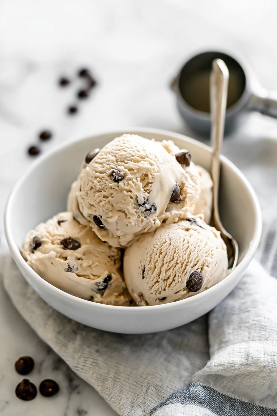 Creamy Cottage Cheese Ice Cream with Chocolate Recipe 8 The image shows a white bowl filled with four scoops of light tan ice cream with dark brown chocolate chips mixed in. The ice cream has a slightly creamy texture and is softly rounded on each scoop, stacked neatly inside the bowl. A metallic spoon with a slim handle rests inside the bowl, pointing outward. The bowl sits on a light cloth with thin stripes, placed on a white marbled surface. In the background, slightly out of focus, there is a metal ice cream scoop and scattered dark chocolate chips. Photo taken with an iphone --ar 2:3 --v 7 - Creamy Cottage Cheese Ice Cream with Chocolate, cottage cheese ice cream, healthy chocolate ice cream, easy homemade ice cream, protein-packed dessert