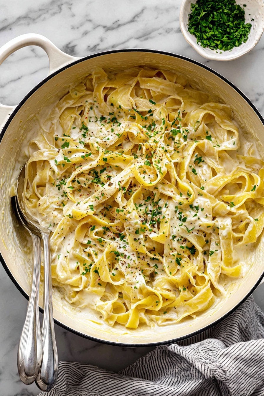 A large white pot filled with creamy yellow pasta ribbons, layered loosely and covered evenly in a smooth, white cream sauce. The pasta is sprinkled with small chopped green herbs and tiny specks of black pepper across the top. Two silver utensils rest inside the pot on the left side, partially covered by the pasta. In the top right corner, there is a small white bowl filled with more green herbs. The pot sits on a white marbled surface with a gray and white striped cloth partially under it. Photo taken with an iphone --ar 2:3 --v 7 - Creamy Lemon Pasta with Parmesan, lemon pasta, easy creamy lemon pasta, pasta with lemon and Parmesan, quick lemon pasta dinner