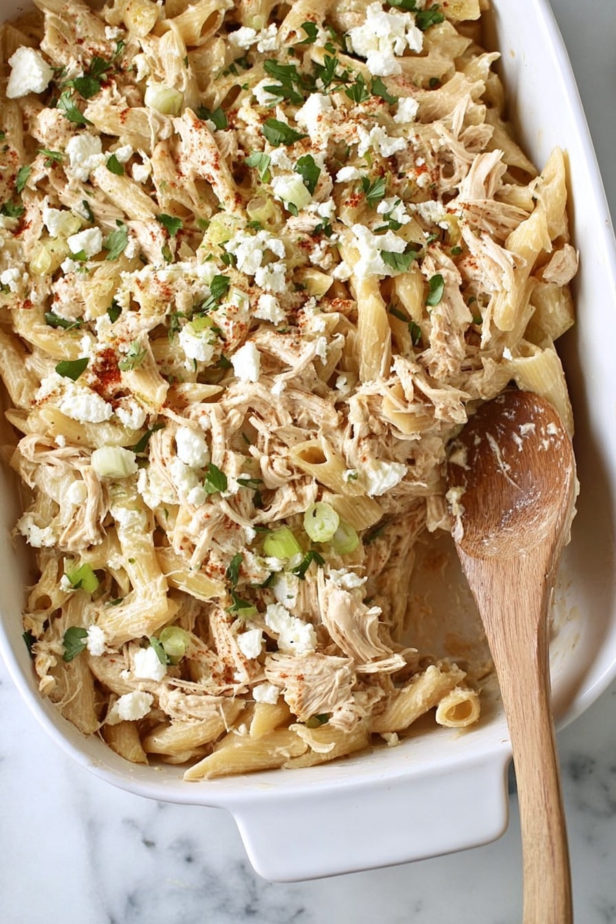 Baked Feta Pasta with Chicken Recipe 6 The image shows a white baking dish filled with a creamy pasta bake. The pasta is light beige and shaped in short tubes mixed with shredded pieces of chicken. Scattered on top are small white clumps of cheese and finely chopped green herbs, adding a fresh contrast. A wooden spoon is resting on the right side of the dish, partly sunk into the pasta. The background is a white marbled surface. photo taken with an iphone --ar 2:3 --v 7 - Baked Feta Pasta with Chicken, baked feta pasta, creamy baked pasta recipe, chicken pasta dish, easy pasta dinner