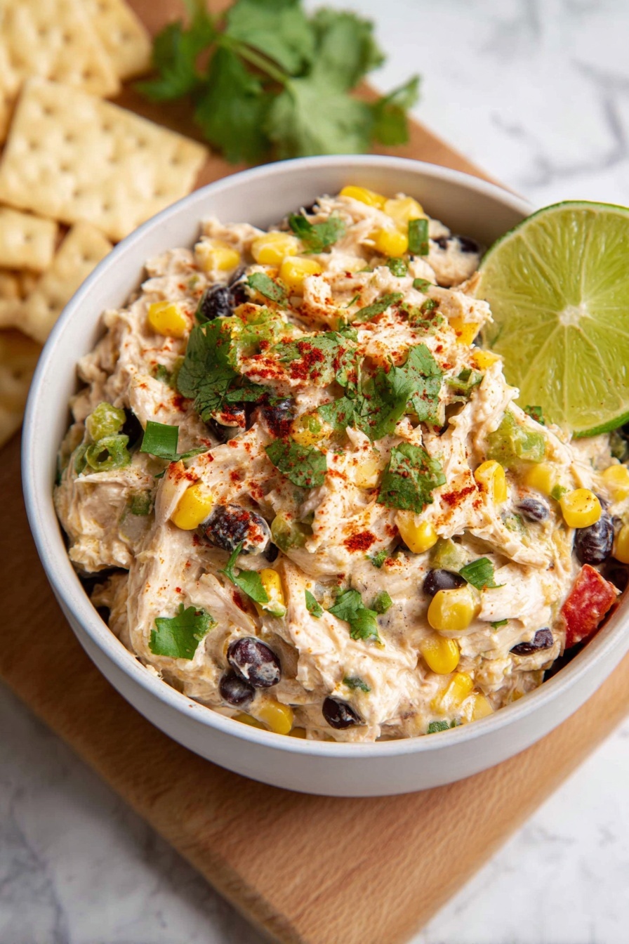 Southwest Chicken Salad Recipe 8 A white bowl is filled with a creamy mixture layered with shredded white chicken meat, yellow corn kernels, whole black beans, and small pieces of green pepper and red tomato. The top is sprinkled with chopped green cilantro leaves and a dusting of red spice powder. A bright green lime wedge rests on the right edge of the bowl. The bowl sits on a wooden board, with some light-colored crackers behind it, all set on a white marbled surface. Photo taken with an iphone --ar 2:3 --v 7 - Southwest Chicken Salad, Southwest Chicken Salad Recipe, flavorful chicken salad, healthy lunch ideas, easy summer salads