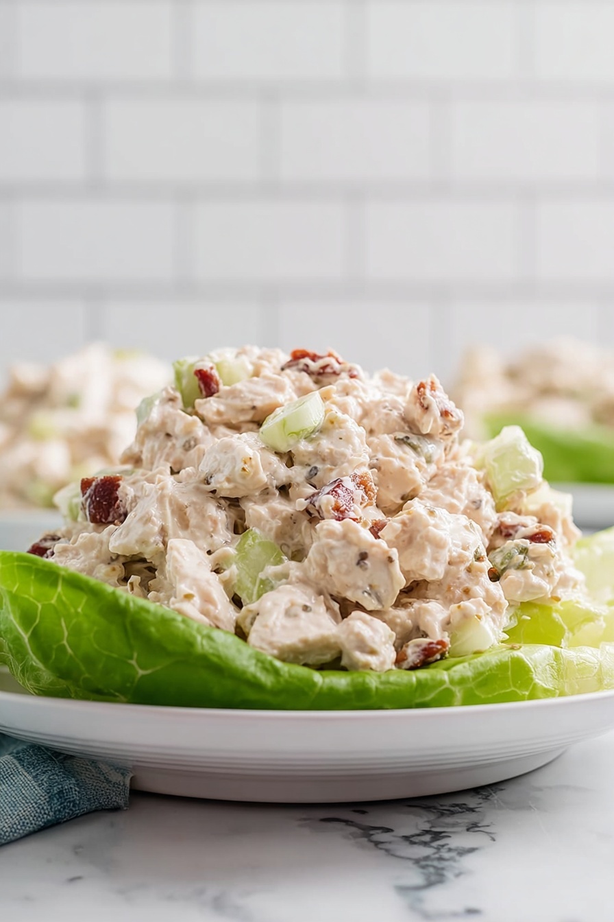 A white plate with a single large green lettuce leaf on the bottom, holding a creamy chicken salad piled high in the center. The chicken salad has a light beige color with visible chunks of white chicken, small bits of celery in pale green, and pieces of reddish-brown nuts or bacon mixed throughout. The creamy texture looks soft and moist, with a few pepper specks scattered in the salad. The background has a white marbled surface and a white tiled wall, giving a clean and bright look. Photo taken with an iphone --ar 2:3 --v 7 - Southern Chicken Salad with Pecans, southern chicken salad, chicken pecan salad, easy southern chicken salad, creamy chicken salad with pecans
